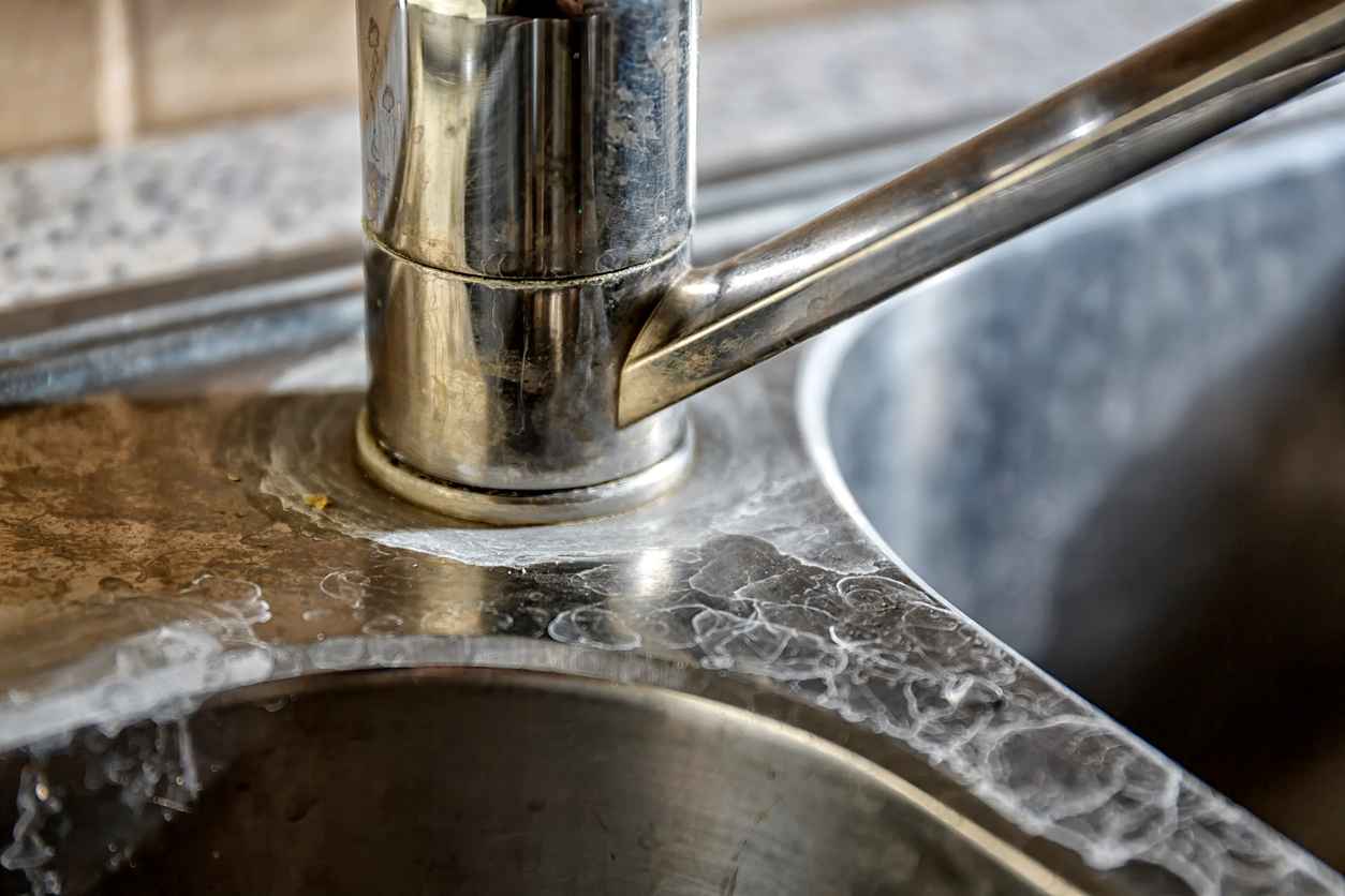 Close-up of a chrome kitchen faucet with a round base, mounted on a marble countertop showing water stains and wear around the sink edge.