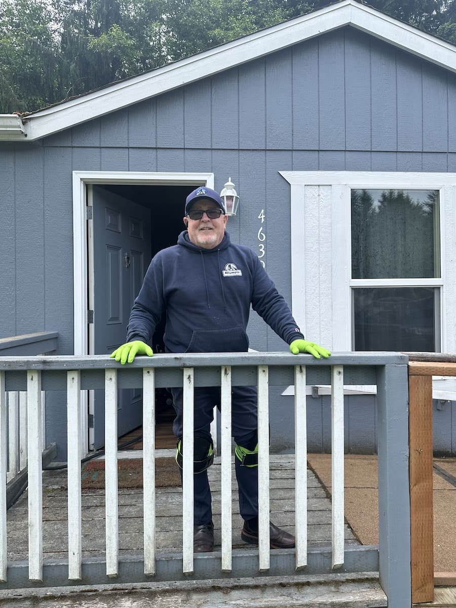Man in a navy hoodie and neon green gloves stands on a wooden porch railing in front of a blue-gray house with the door open behind him.