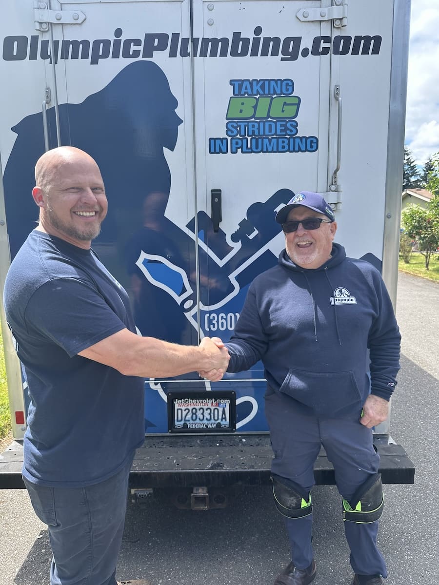Two men shake hands in front of a plumbing truck with OlympicPlumbing branding and slogan 'Taking Big Strides in Plumbing'.