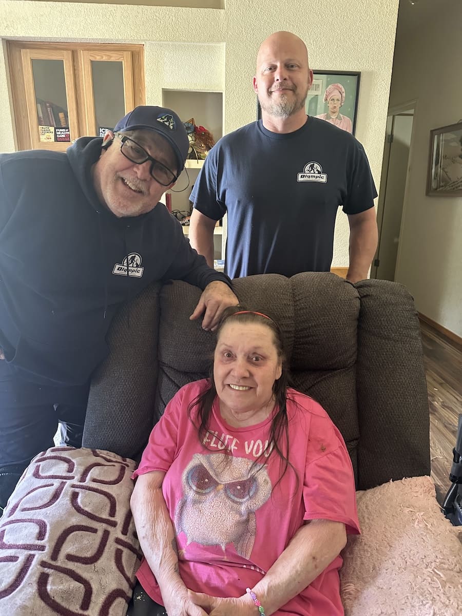Two male caregivers in navy shirts stand beside an elderly woman sitting on a brown recliner in a living room, smiling for the photo.