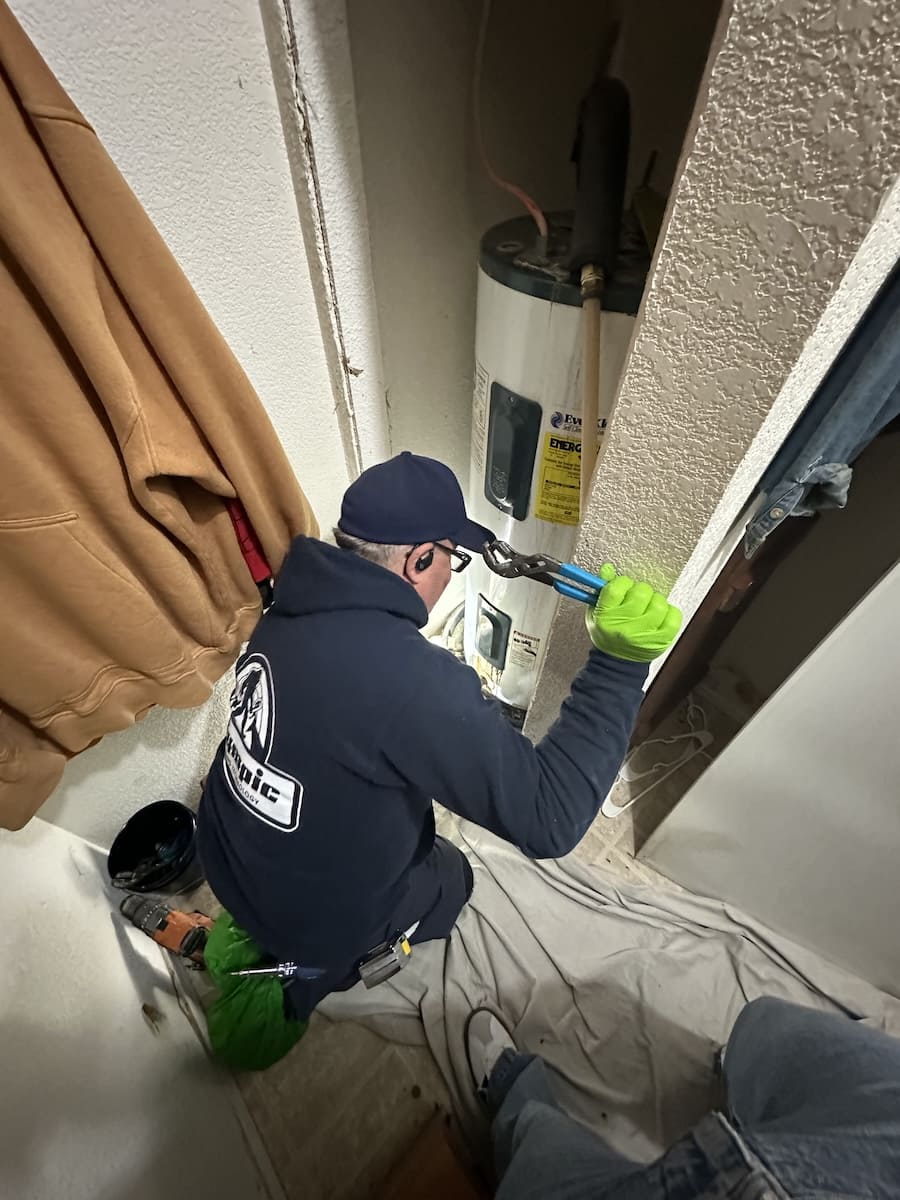 Plumber kneeling in a closet, using pliers on a water heater with bright green gloves.