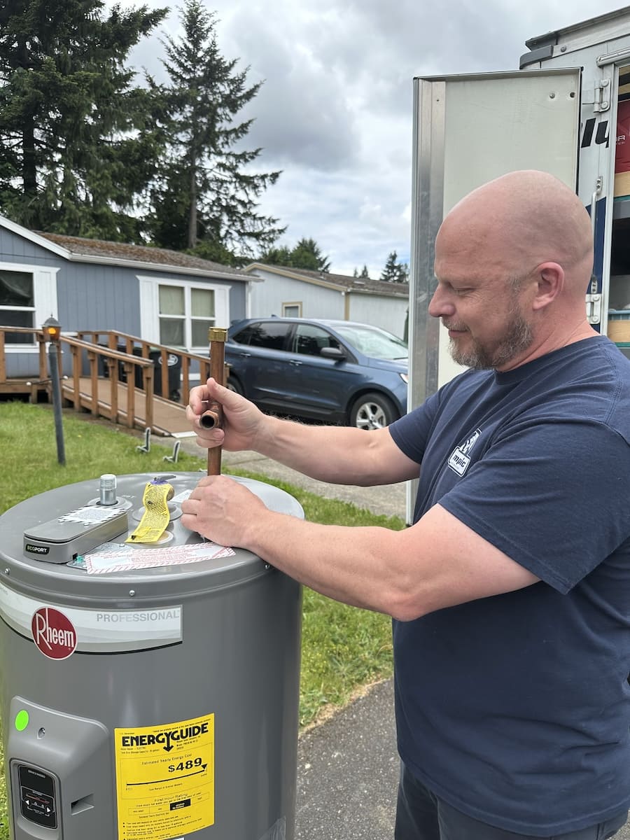 Man in a navy T-shirt working on a Rheem professional water heater outdoors, using a pipe wrench near the top access point with a truck in the background.