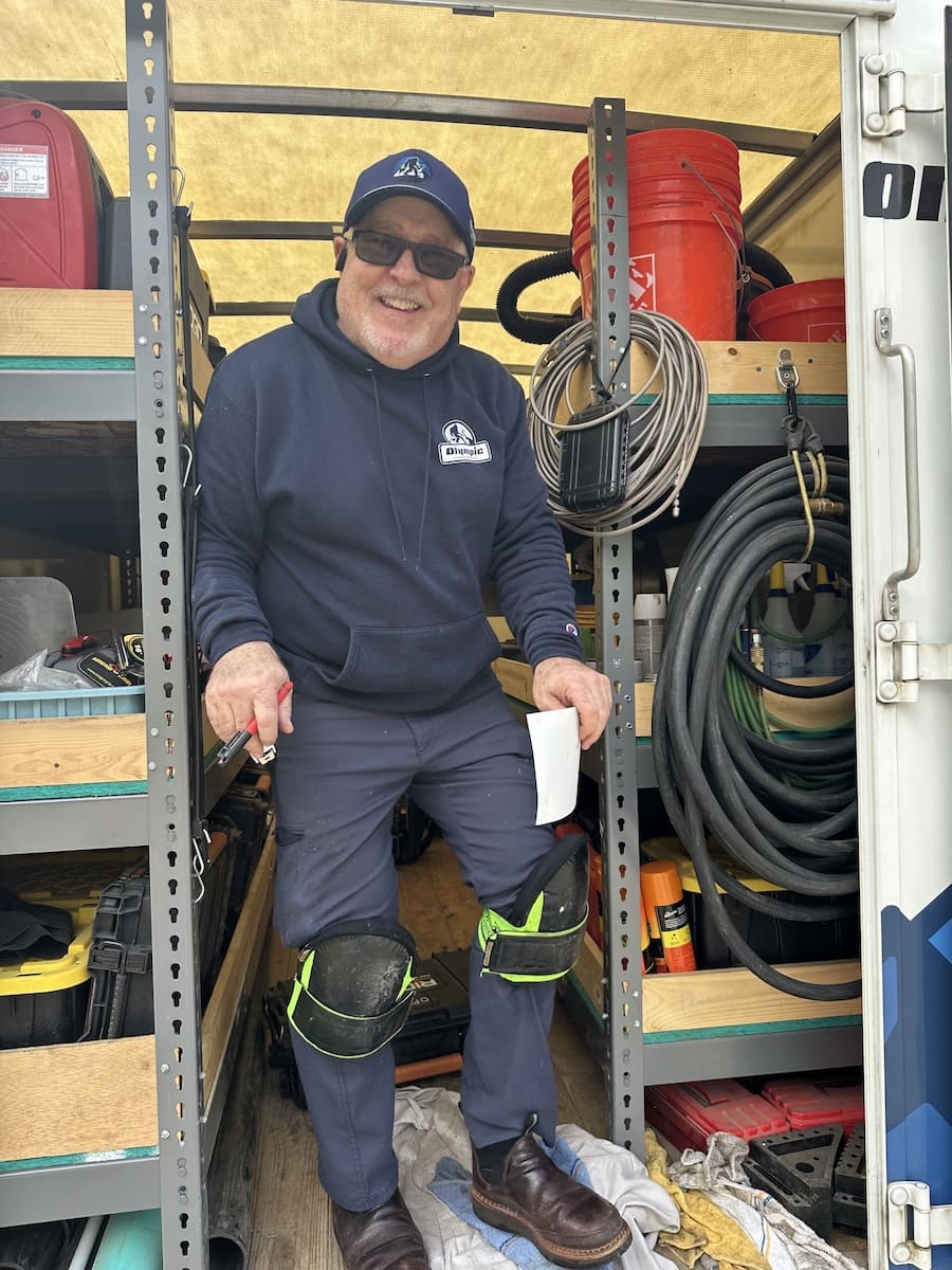 Man in a navy hoodie and cap standing inside a work van with shelves of tools, knee pads, and coiled cables, smiling at the camera while holding a marker and a small sheet of paper.