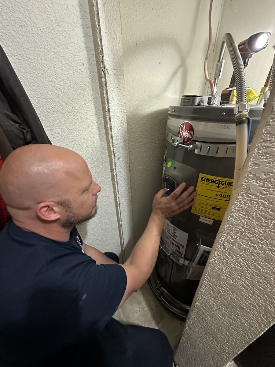 Technician kneeling in a narrow utility closet, adjusting the control panel on a Rheem water heater.