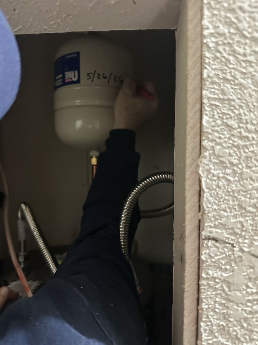 Plumber's hand writing the date 5/26/24 on a white expansion tank inside a utility cabinet