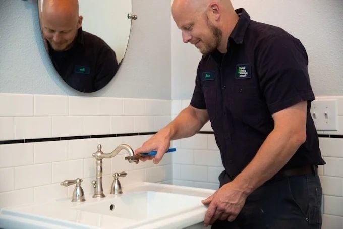 Male plumber in a dark uniform tightens a blue-handled faucet at a white bathroom sink, reflected in a round mirror.