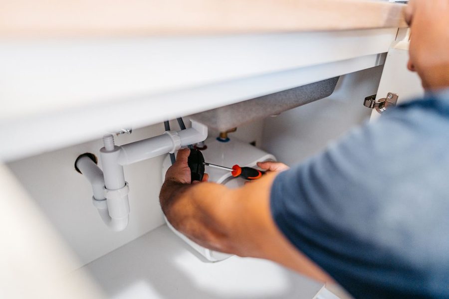 Plumber under a kitchen sink tightening pipes with a screwdriver.