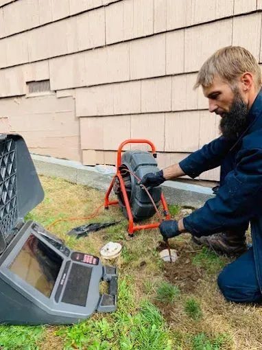 Man kneeling on grass beside a wall, using a handheld tester connected to a portable diagnostic device and red test stand for an outdoor soil/drainage check.
