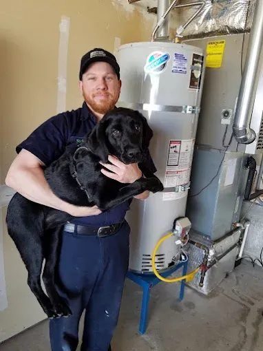Technician in navy work uniform holding a black dog in a utility room with a water heater nearby.